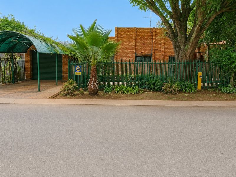 Front view of a brick house featuring a green metal carport, secure perimeter fencing, and established garden elements.