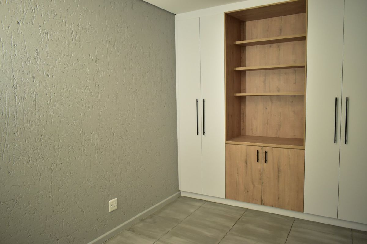A clean, empty bedroom featuring modern built-in cupboards with integrated shelving, neutral grey textured walls, and tiled flooring.