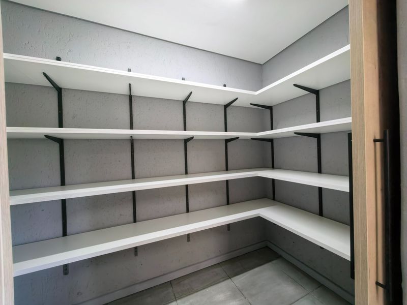 A clean, modern walk-in pantry or scullery area featuring extensive built-in white shelving with black support brackets and neutral tiled flooring.