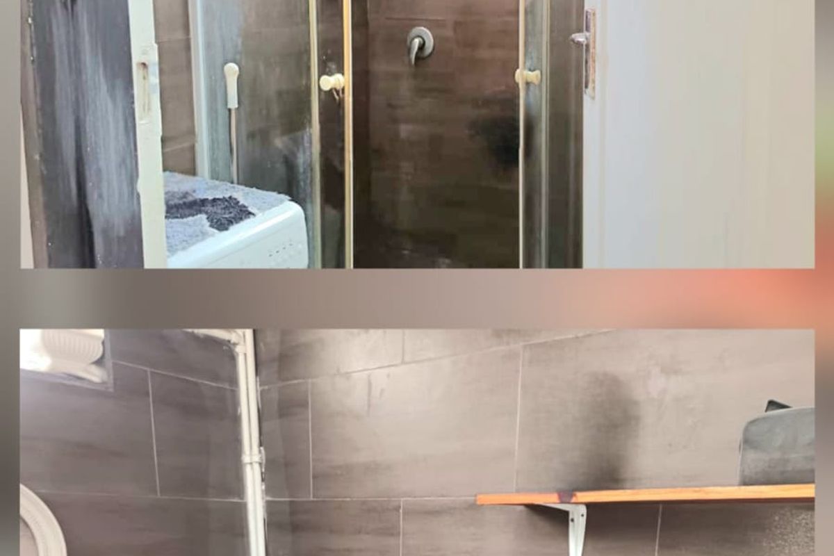 A bathroom featuring dark wall tiling, a glass shower enclosure, a standard toilet, and wall-mounted shelving with visible plumbing.