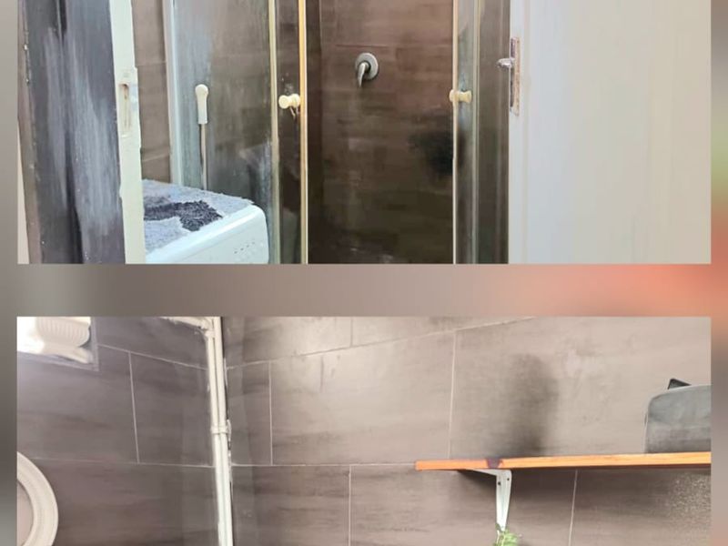 A bathroom featuring dark wall tiling, a glass shower enclosure, a standard toilet, and wall-mounted shelving with visible plumbing.