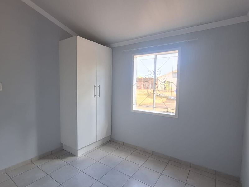 A clean, empty bedroom featuring neutral tiled flooring, a built-in white wardrobe, and a window fitted with decorative security bars.