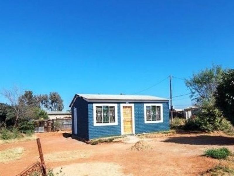 A detached single-story structure featuring a blue brick facade, corrugated metal roof, and security-fitted windows, situated on an unpaved plot.