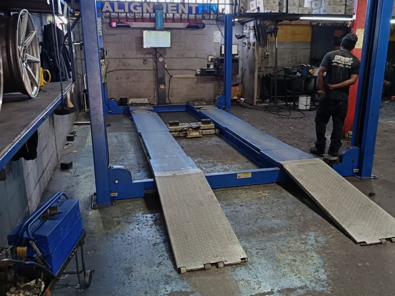 Commercial automotive workshop featuring a vehicle alignment lift, industrial steel shelving, and exposed structural elements with concrete flooring.