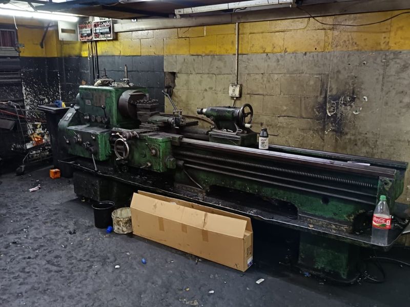 Industrial workshop space featuring a heavy-duty metal lathe, overhead storage shelving, and exposed concrete block walls with fluorescent lighting.
