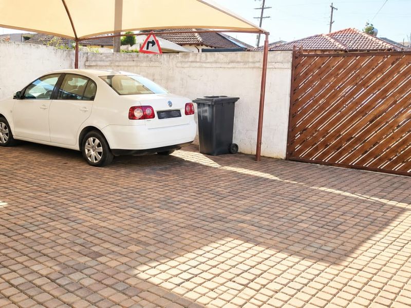 A paved outdoor parking area featuring a shade sail, perimeter wall, and a wooden gate.