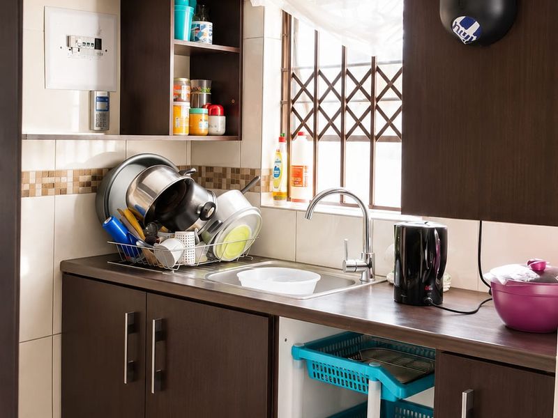 A compact scullery area featuring dark wood-look cabinetry, a stainless steel sink, and tiled splashback with a window security gate.