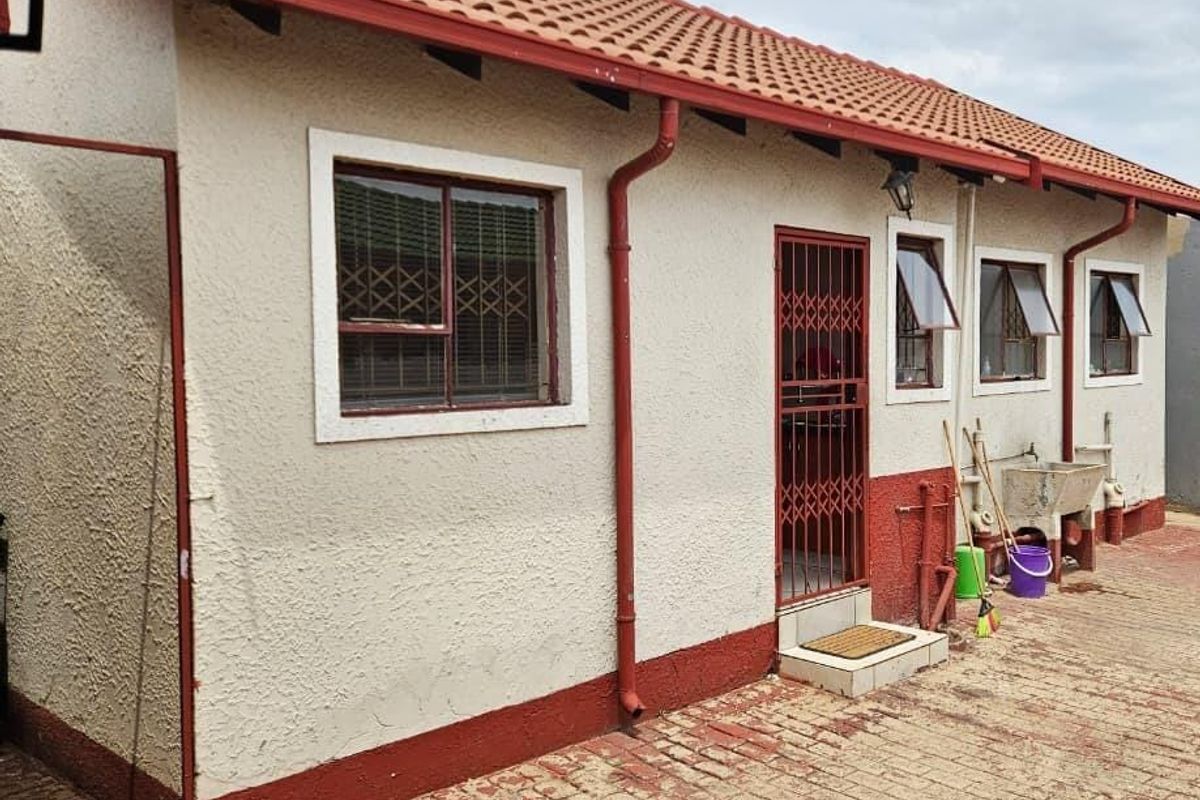 Rear exterior view of a house featuring a terracotta tiled roof, textured walls, security gates, and a paved courtyard area with an external utility sink.
