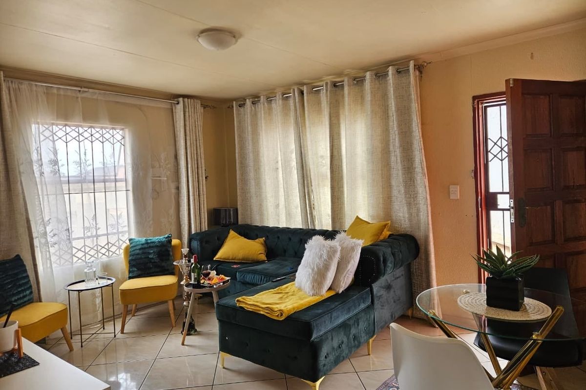 A comfortable living room featuring tiled flooring, natural light through large windows, and direct access to the exterior via a security door.