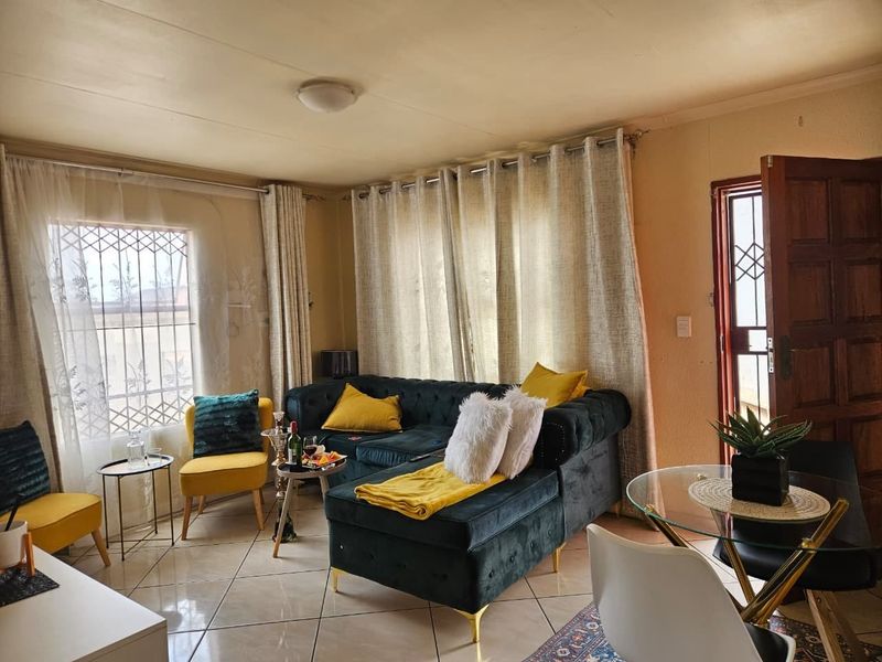 A comfortable living room featuring tiled flooring, natural light through large windows, and direct access to the exterior via a security door.