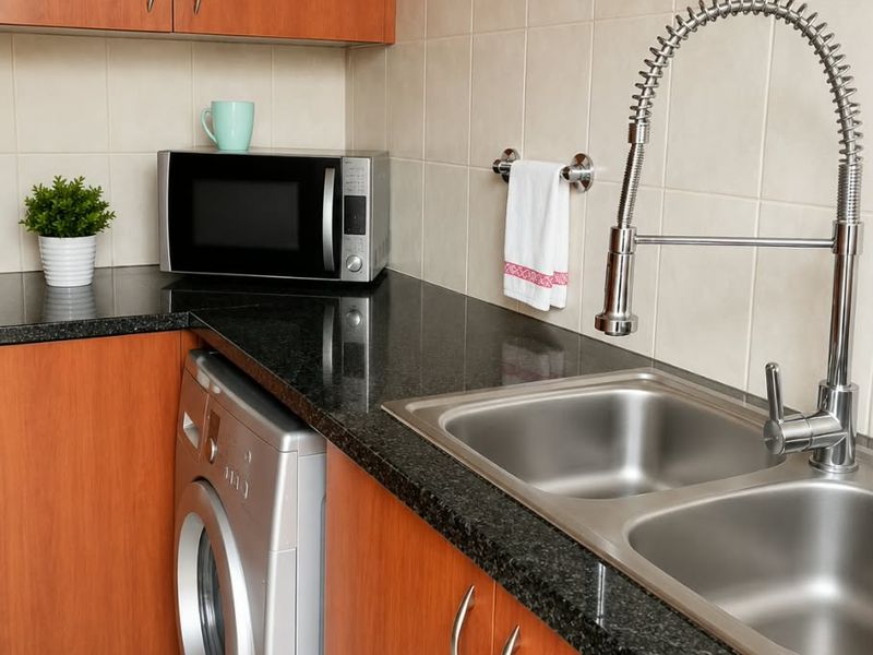 A functional kitchen area featuring wood-finish cabinetry, dark granite countertops, a double stainless steel sink with a modern gooseneck faucet, and tiled backsplash.