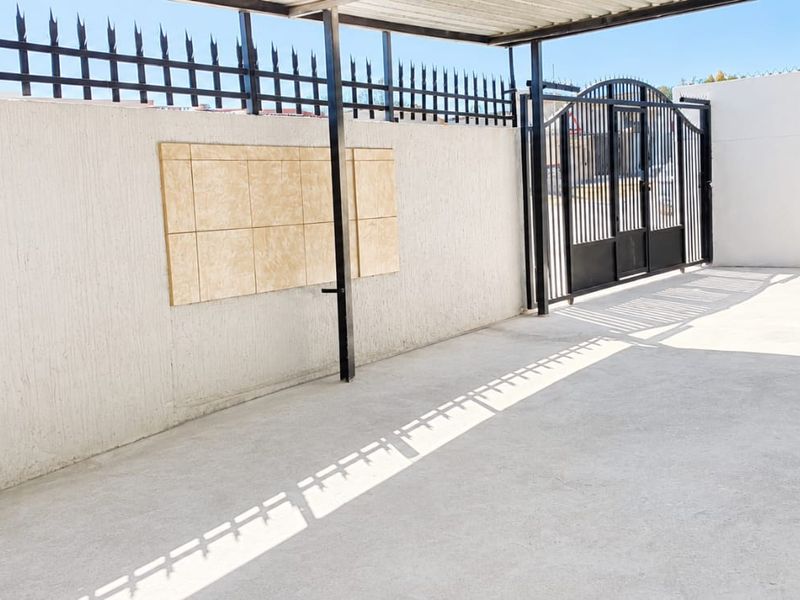 A clean, paved outdoor carport area featuring a secure metal gate, perimeter fencing, and a covered roof structure.