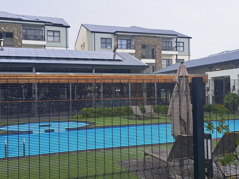 A well-maintained communal swimming pool area within an apartment complex, featuring modern multi-story buildings, solar panels, and secure perimeter fencing.