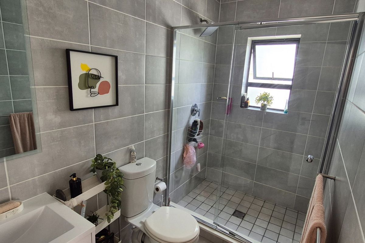 Modern bathroom featuring grey wall tiles, a walk-in glass shower, vanity basin, and toilet, finished with contemporary fixtures and a casement window.