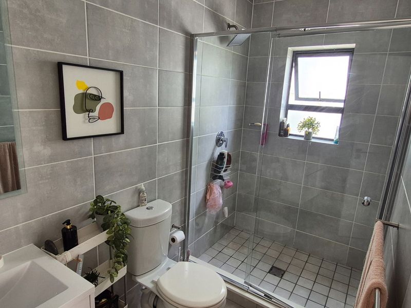 Modern bathroom featuring grey wall tiles, a walk-in glass shower, vanity basin, and toilet, finished with contemporary fixtures and a casement window.