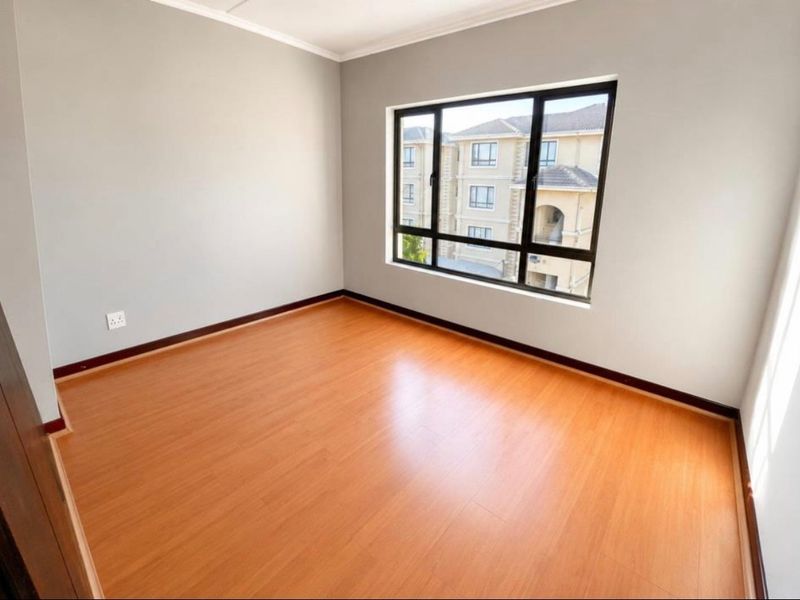 A bright, empty bedroom featuring laminate flooring, neutral grey walls, and large aluminium-framed windows providing natural light.