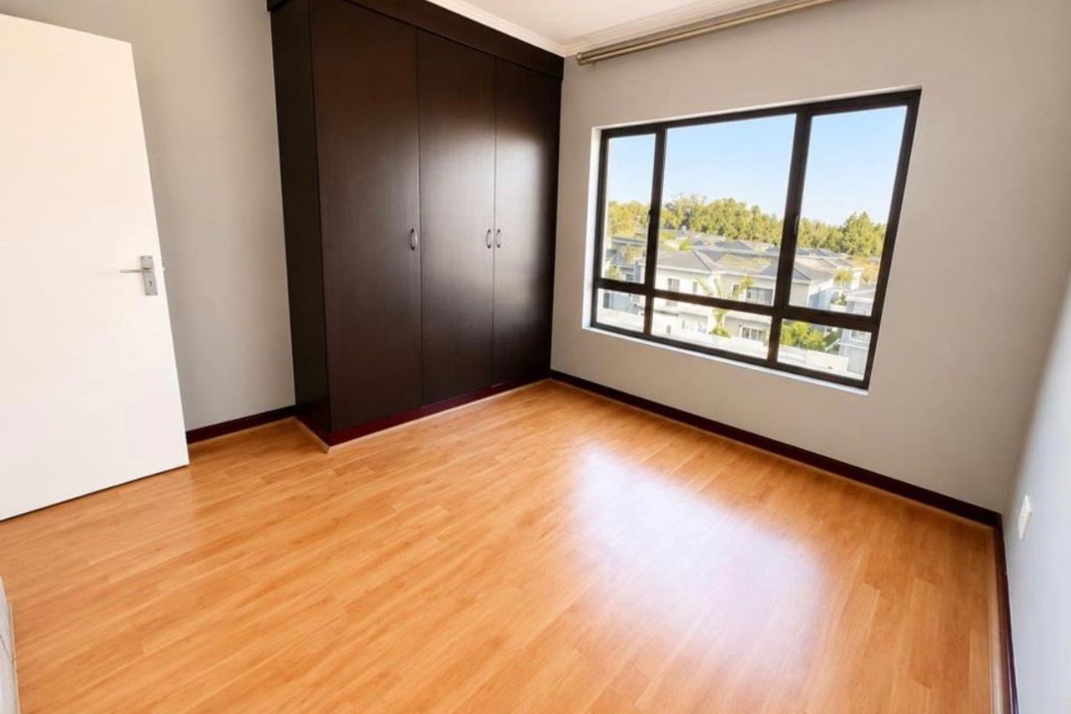 A bright, empty bedroom featuring laminate flooring, built-in dark wood cupboards, and a large window with a view of the surrounding neighborhood.