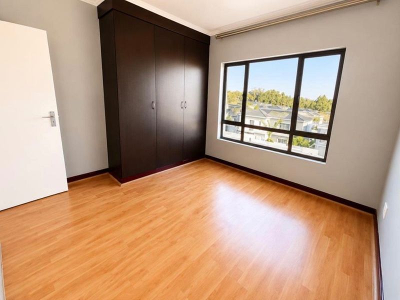 A bright, empty bedroom featuring laminate flooring, built-in dark wood cupboards, and a large window with a view of the surrounding neighborhood.