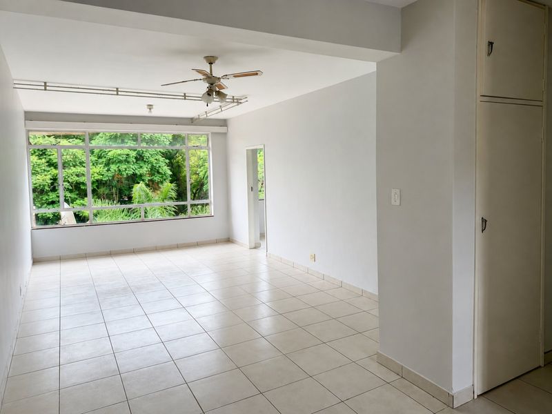 A bright, empty living area featuring tiled floors, a ceiling fan, and a large window with a pleasant garden view.