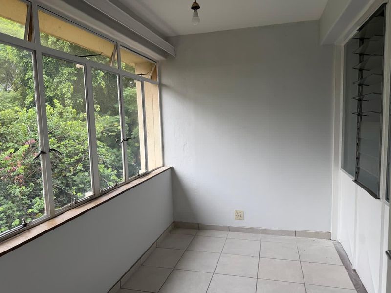 An empty, enclosed balcony featuring tiled flooring and large windows with views of surrounding greenery, offering a versatile space for a sunroom or study.