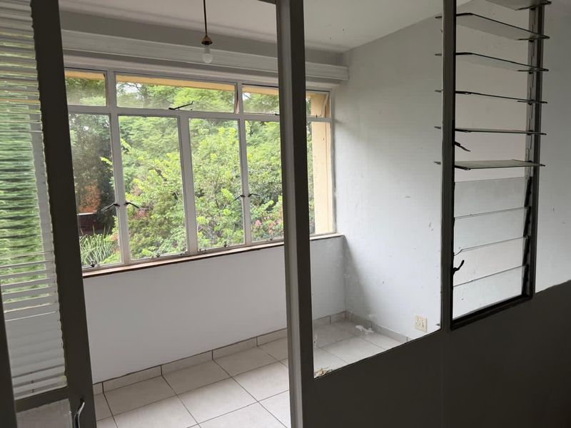 An enclosed balcony or sunroom featuring tiled flooring, large aluminium-framed windows, and louvered ventilation panels, offering a view of the surrounding greenery.