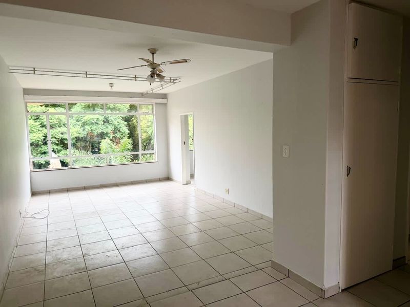 An empty, open-plan living area featuring tiled flooring, a ceiling fan, and a large window providing natural light and garden views.