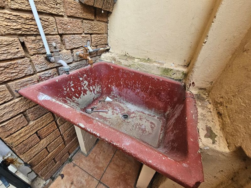 An outdoor utility area featuring a worn, red-painted concrete sink with exposed plumbing fixtures against a brick and plastered wall.