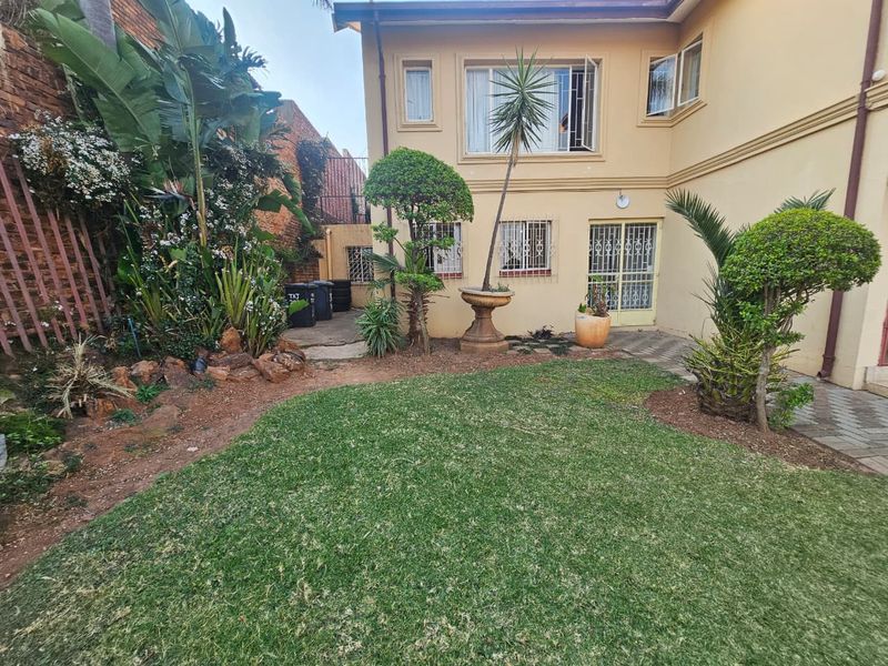 A rear view of the property featuring a well-maintained lawn, landscaped garden beds, and paved access to the building entrance.
