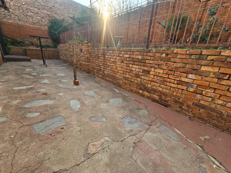 A rear outdoor area featuring a brick retaining wall, metal fencing, and weathered crazy paving, offering potential for landscaping and patio development.