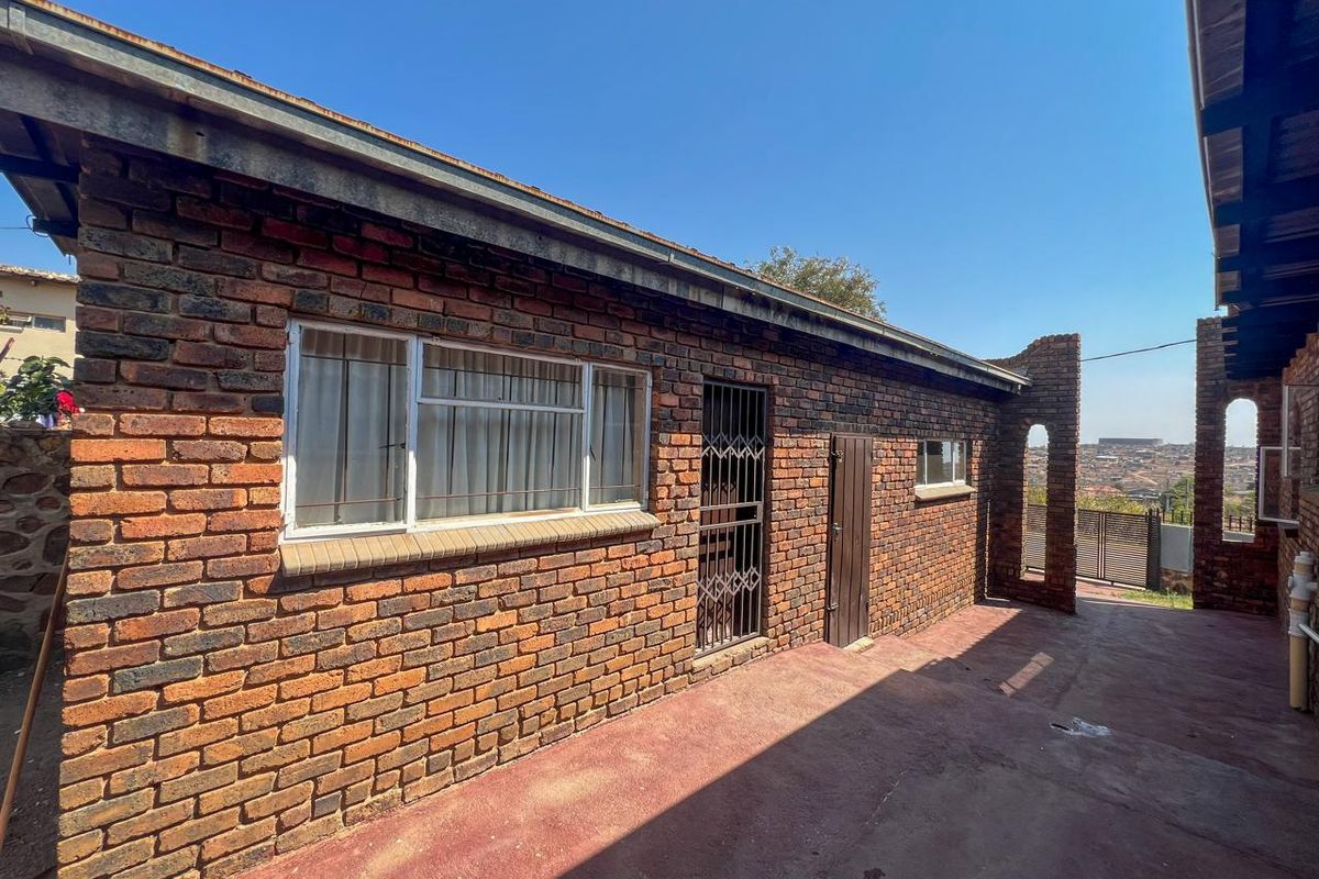 Rear exterior view of a face brick house featuring a concrete patio, metal security gate, and aluminum window frames under a clear sky.