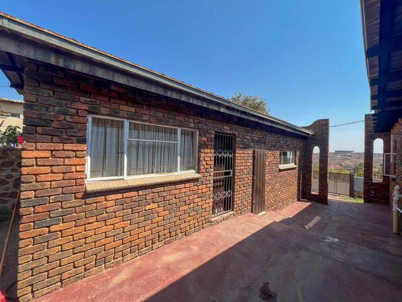 Rear exterior view of a face brick house featuring a concrete patio, metal security gate, and aluminum window frames under a clear sky.