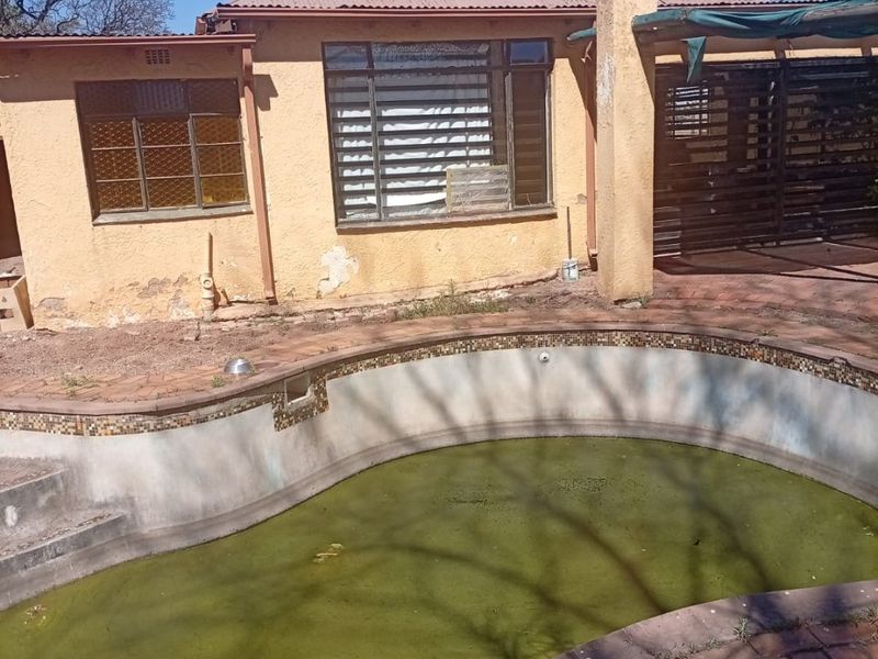 Rear exterior view featuring a swimming pool with stagnant water, brick paving, and a covered patio area attached to the main house structure.