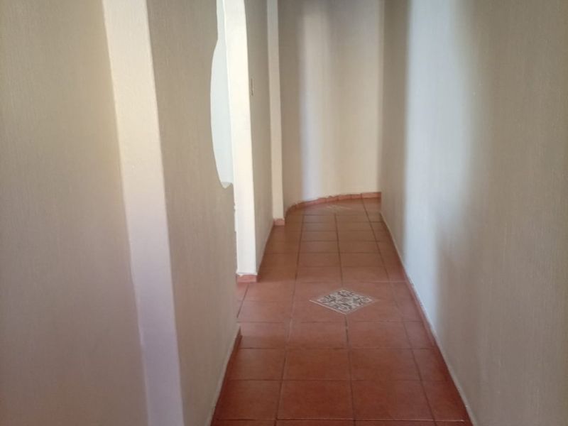 A long, empty hallway featuring terracotta floor tiles with decorative inserts, recessed ceiling lighting, and a unique arched wall opening.