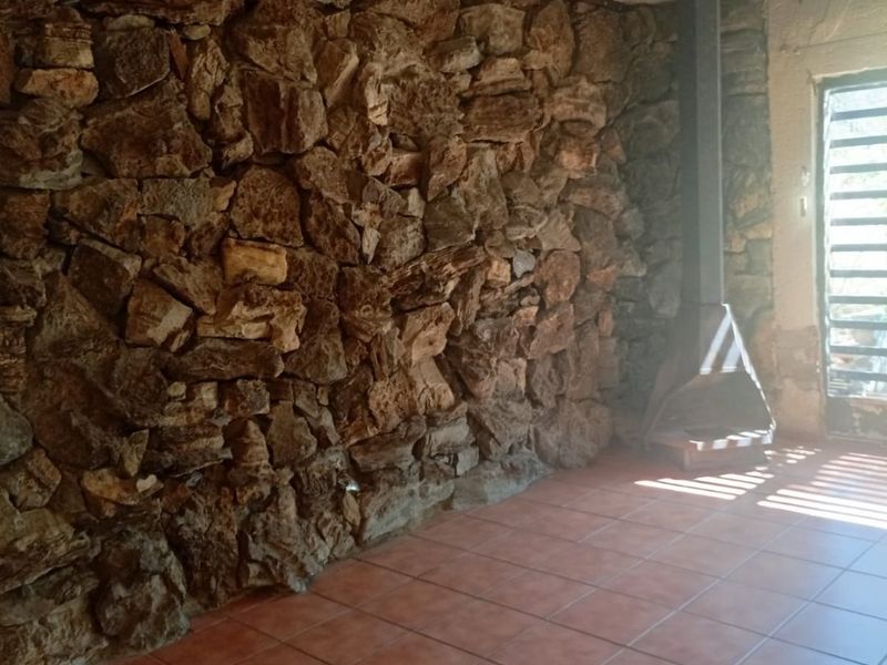 An empty living area featuring a prominent stone feature wall, a freestanding fireplace, ceramic tiled flooring, and exposed ceiling beams.