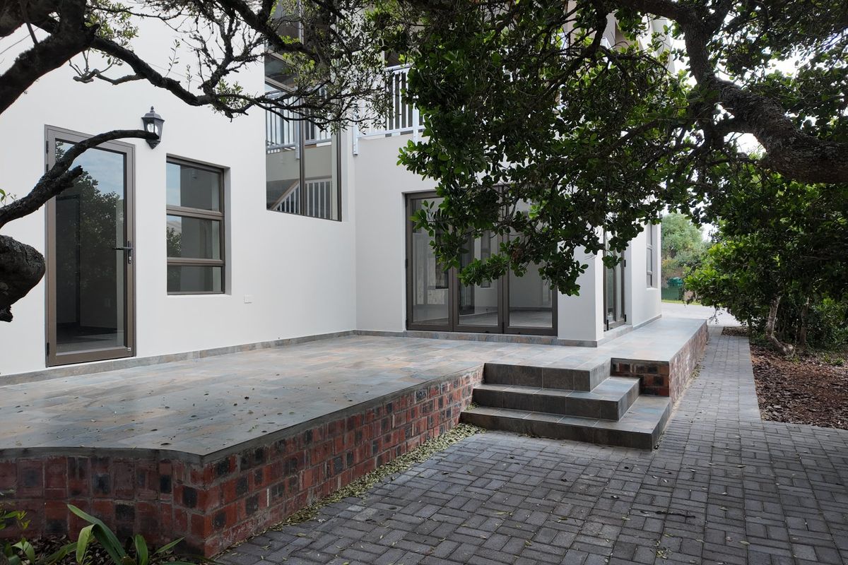 A well-maintained rear exterior featuring a tiled patio with a brick retaining wall, paved pathways, and modern aluminium-framed doors and windows.