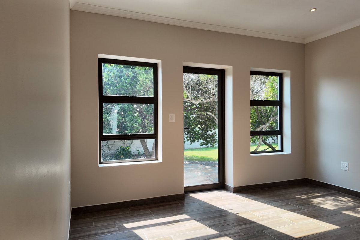 A bright, empty living area featuring modern tiled flooring, neutral walls, and large aluminium-framed windows and a door providing access to the garden.