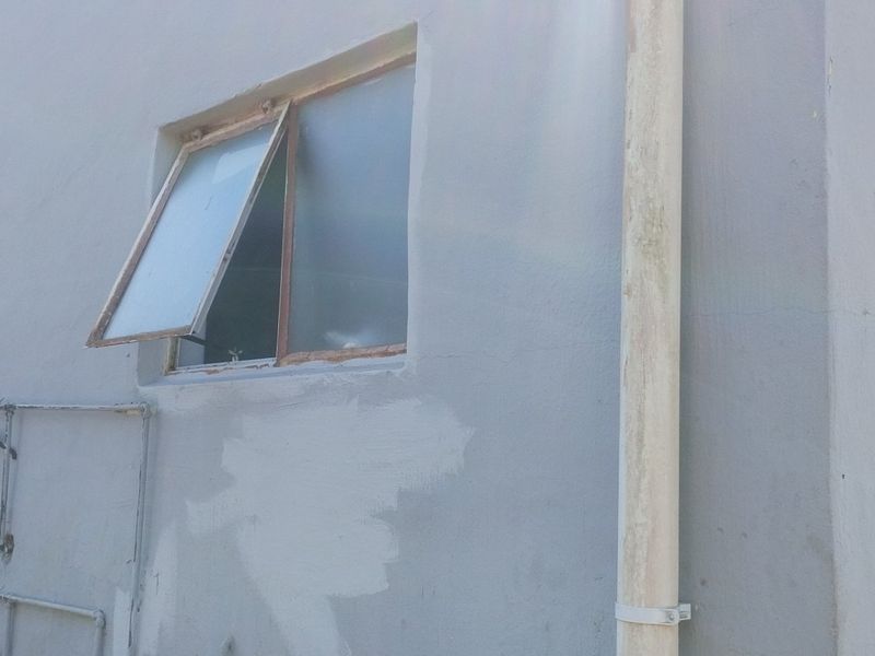 A view of the exterior rear wall showing a casement window, a weathered downpipe, and exposed plumbing fixtures against an unfinished painted surface.