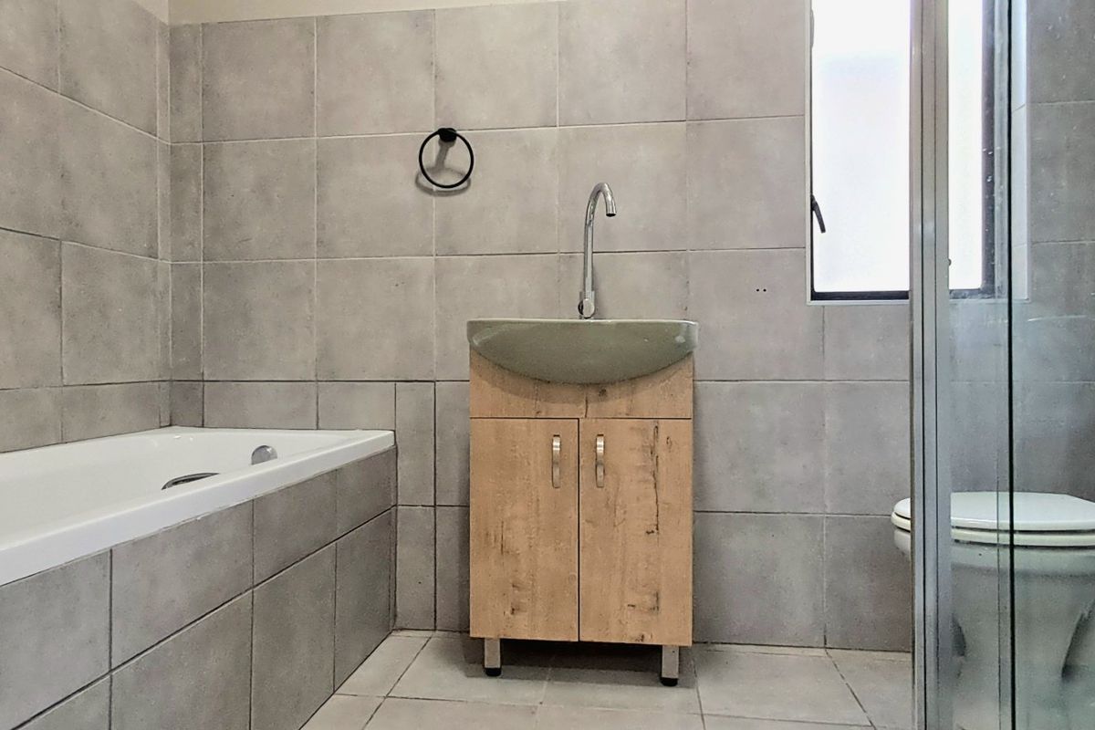 A modern bathroom featuring a built-in bathtub, a vanity cabinet with a basin, a toilet, and a glass-enclosed shower with neutral grey wall tiles.