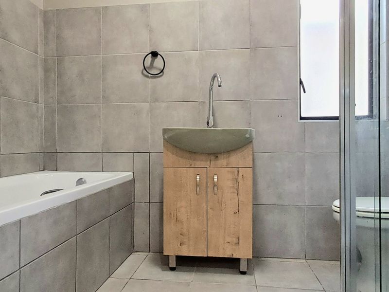 A modern bathroom featuring a built-in bathtub, a vanity cabinet with a basin, a toilet, and a glass-enclosed shower with neutral grey wall tiles.