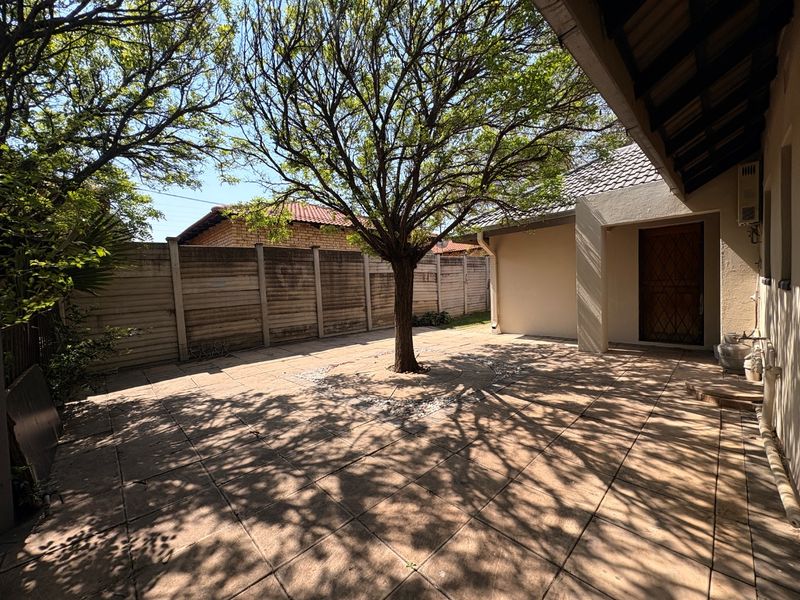 A paved outdoor patio area featuring a central tree, precast concrete boundary walls, and direct access to the home via a security gate.