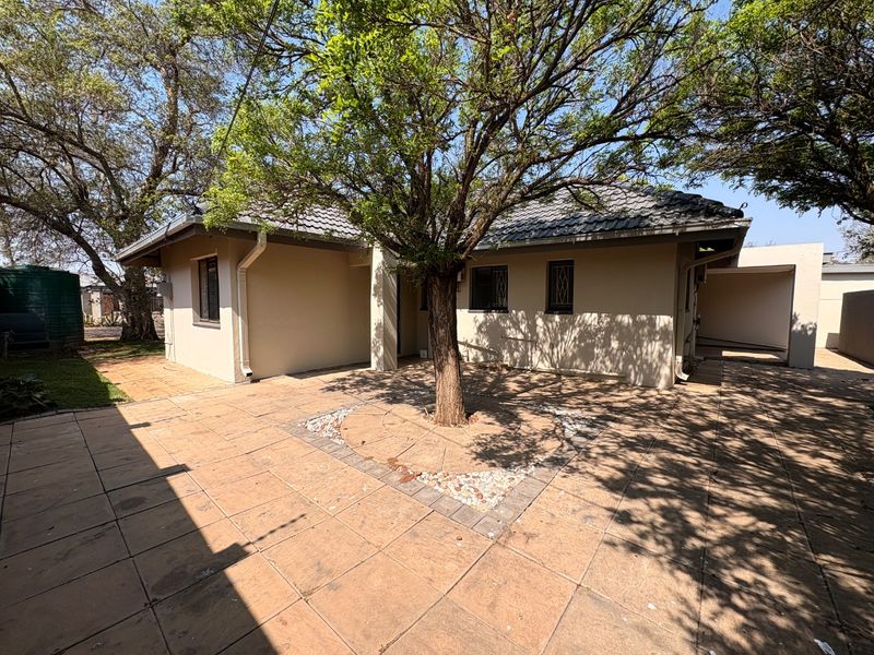 A well-maintained rear exterior view featuring a paved courtyard, mature tree, and tiled patio area with security bars on the windows.