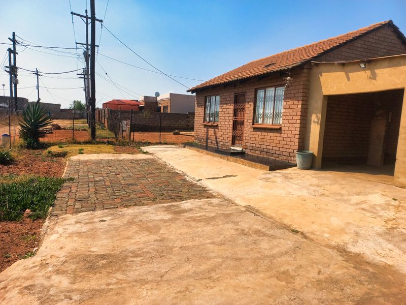 Front view of a single-story brick house featuring a tiled roof, security gate, and a paved driveway with an attached carport area.
