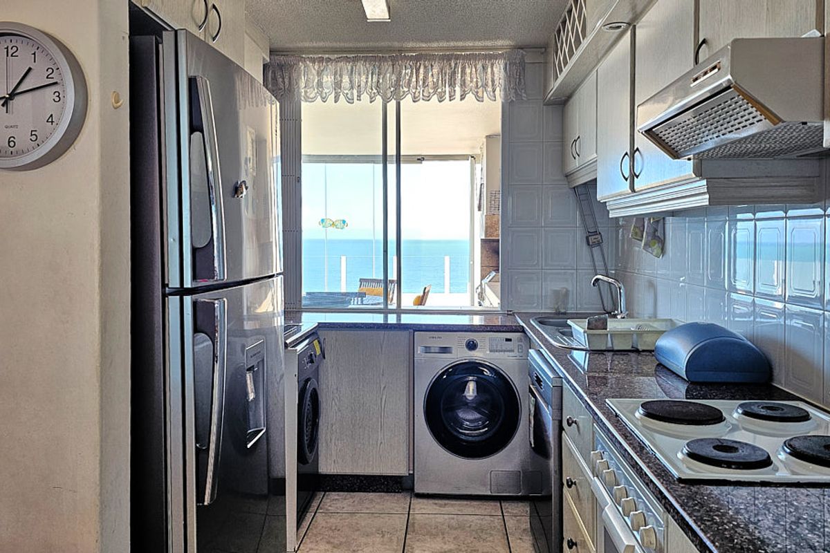 Compact galley kitchen featuring granite countertops, tiled backsplash, built-in hob, extractor fan, and integrated laundry appliances, with direct access to a balcony and ocean view.