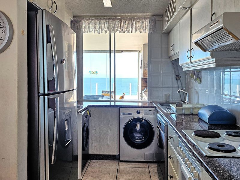 Compact galley kitchen featuring granite countertops, tiled backsplash, built-in hob, extractor fan, and integrated laundry appliances, with direct access to a balcony and ocean view.