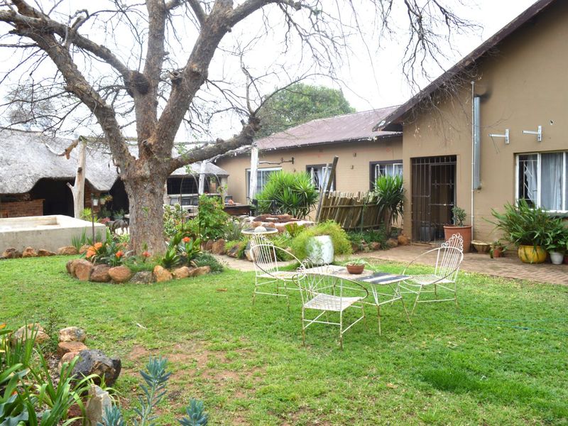 A rear garden view featuring a lawn, mature tree, paved patio area, and a secondary thatched structure in the background.