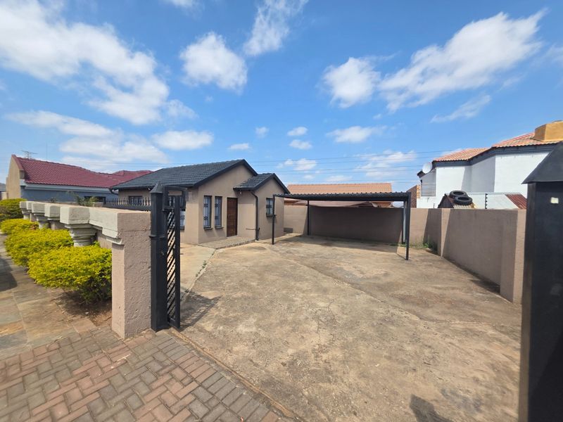 Front view of the property featuring a concrete driveway, covered carport, and secure boundary wall with metal gate access.