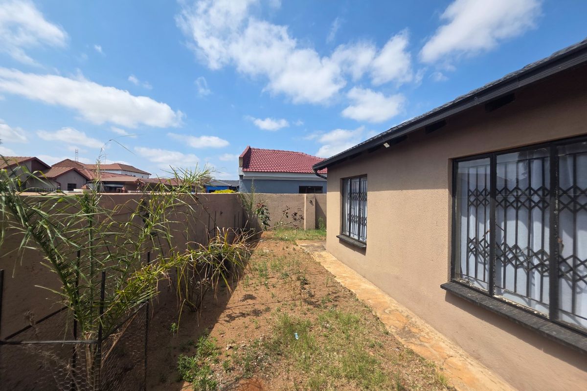 A rear exterior view of the property featuring a paved walkway, security bars on windows, and a perimeter wall enclosing the garden area.