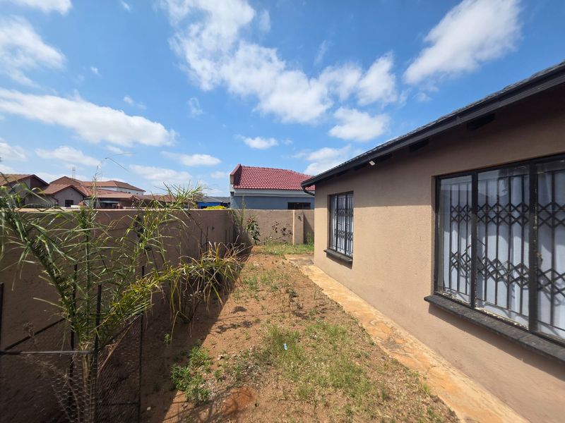 A rear exterior view of the property featuring a paved walkway, security bars on windows, and a perimeter wall enclosing the garden area.