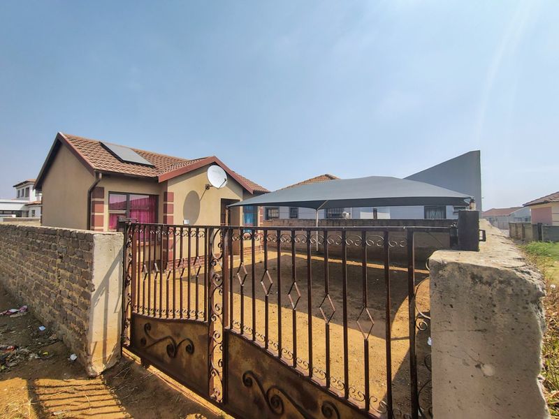 Front view of a residential house featuring a tiled roof with solar water heater, enclosed by a brick boundary wall and wrought iron gate.