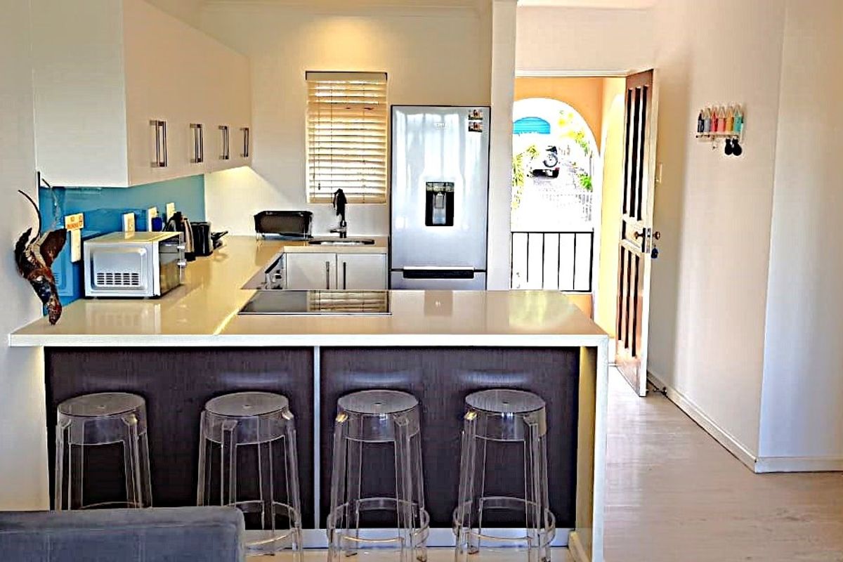 Modern kitchen featuring sleek white cabinetry, stone countertops, an integrated electric hob, and a breakfast bar, leading to an outdoor balcony area.